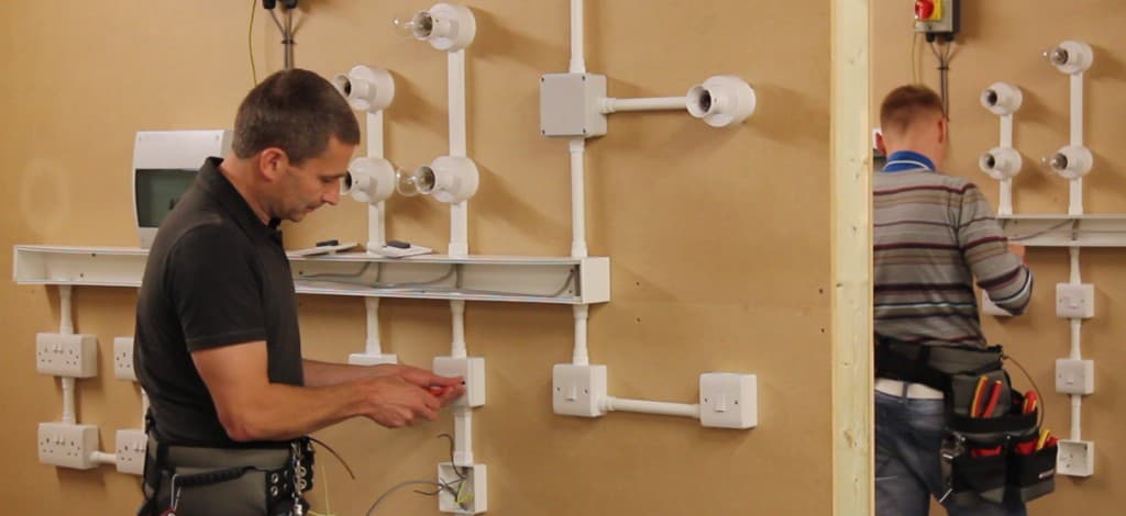 Two electricians installing wiring and fixtures on a training board